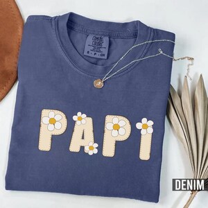 May include: A navy blue t-shirt with the word "PAPI" in white letters with a daisy design. The daisies have white petals and yellow centers.