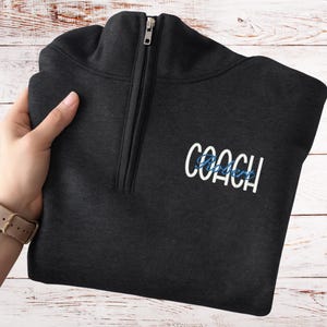 May include: A dark gray quarter-zip sweatshirt with the word "COACH" embroidered in white and a blue script name underneath. The sweatshirt has a silver zipper and is folded. The background is a light wood grain.