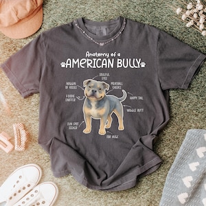 May include: Charcoal grey t-shirt with a diagram of an American Bully, detailing body parts such as "Soulful Eyes" and "Happy Tail." The text "Anatomy of a AMERICAN BULLY" is displayed. The shirt is surrounded by accessories.
