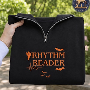 May include: A black quarter-zip sweatshirt featuring orange embroidery that reads RHYTHM READER. The design includes a small ghost, a heartbeat line, and three flying bats arranged around the text. The garment has a silver metal zipper.