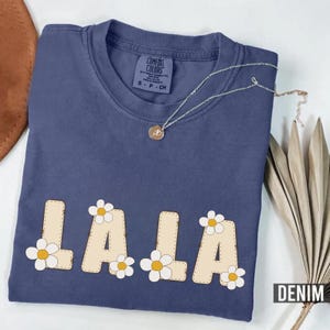 Puede incluir: Una camiseta Comfort Colors azul denim con la palabra "LALA" en una fuente beige cosida, decorada con flores de margarita blancas y amarillas. Un collar plateado con un colgante está sobre la camiseta. La palabra "DENIM" está en la esquina inferior derecha.