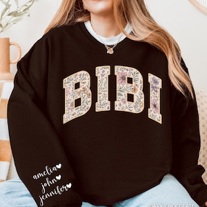 Puede incluir: Sudadera de color chocolate oscuro con el nombre "BIBI" en un diseño floral en el pecho. La manga izquierda presenta los nombres "amelia", "john" y "jennifer". El texto "DARK CHOCOLATE" está en la parte inferior derecha.