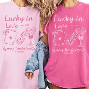 Puede incluir: Dos camisetas rosas con el texto "Lucky in Love est. 2023" y "Graces Bachelorette Los Cabos, Mexico". El diseño incluye naipes, dados y una copa de cóctel. Una camiseta es rosa claro, la otra es rosa intenso.