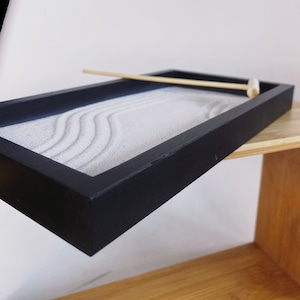 May include: A black wooden tray with a white sand garden inside. The tray has a wooden rake for creating patterns in the sand.