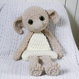 Pode incluir: Um brinquedo de pel&uacute;cia de croch&ecirc; em forma de elfo dom&eacute;stico, com grandes olhos verdes e um vestido branco de croch&ecirc;. O brinquedo est&aacute; sentado em um cobertor branco de croch&ecirc;.