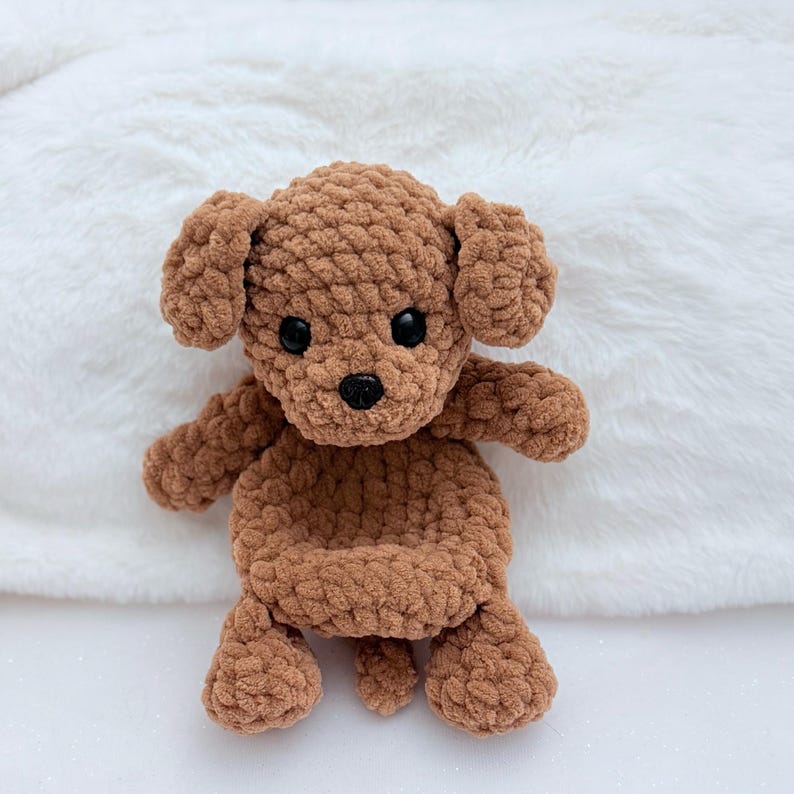 Puede incluir: Un perro de peluche de crochet marr&oacute;n con ojos negros y nariz negra. El perro est&aacute; sentado sobre una superficie blanca y esponjosa.