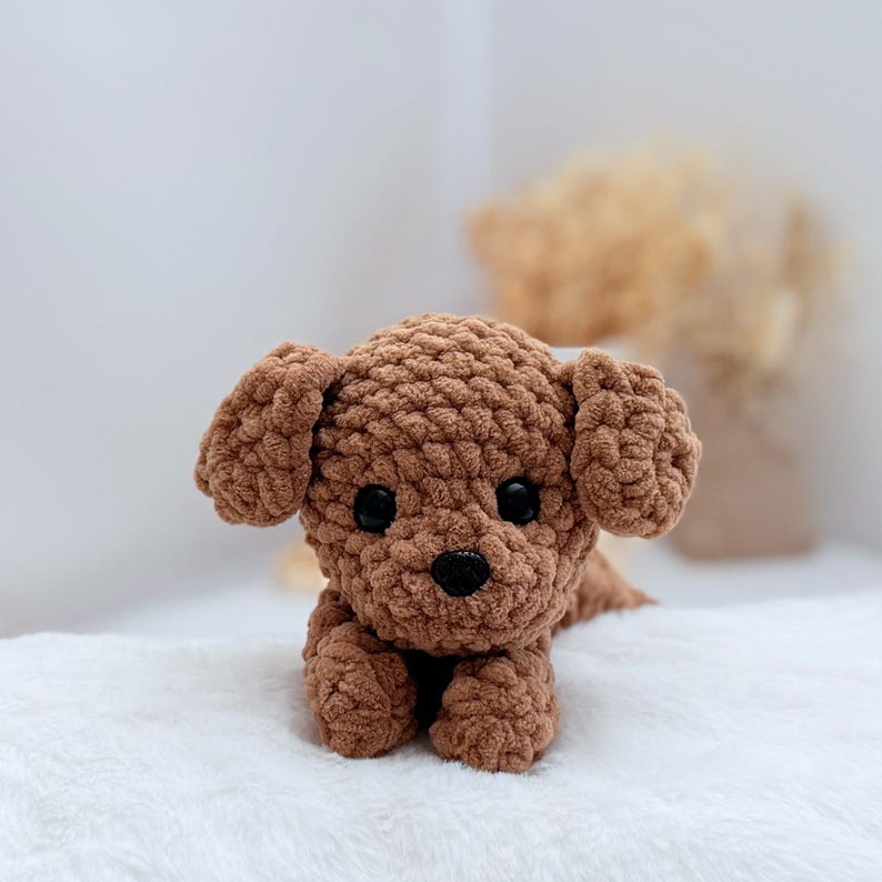 Puede incluir: Un perro de peluche de crochet marr&oacute;n con ojos negros y nariz negra. El perro est&aacute; acostado con las patas delante.