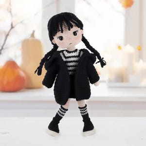 May include: A handmade crochet doll with black yarn hair styled in two braids, wearing a black hooded cardigan over a black and white striped shirt, a black skirt, and black and white striped knee-high socks with white shoes on a light surface.