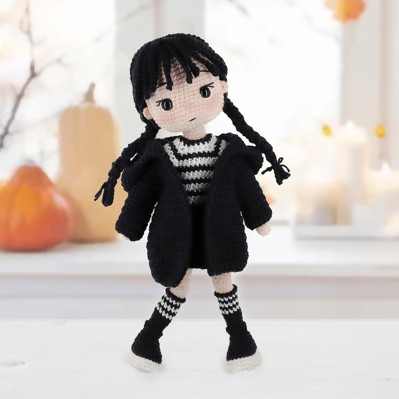 May include: A handmade crochet doll with black yarn hair styled in two braids, wearing a black hooded cardigan over a black and white striped shirt, a black skirt, and black and white striped knee-high socks with white shoes on a light surface.