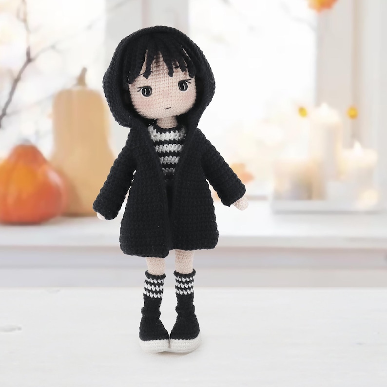 May include: A handmade crochet doll featuring a pale skin tone, black hair, and dark eyes. The doll wears a black hooded cardigan over a black and white striped shirt, paired with black and white striped socks and black shoes with white soles.