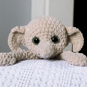 Pode incluir: Um elefante de pel&uacute;cia de croch&ecirc; com grandes olhos verdes. O elefante est&aacute; deitado em um cobertor branco de croch&ecirc;.