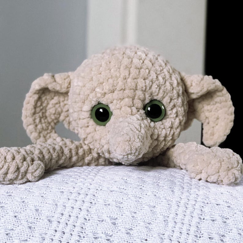 Pode incluir: Um elefante de pel&uacute;cia de croch&ecirc; com grandes olhos verdes. O elefante est&aacute; deitado em um cobertor branco de croch&ecirc;.