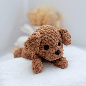 Puede incluir: Un perro de peluche de crochet marr&oacute;n con ojos negros y nariz negra. El perro est&aacute; acostado sobre una superficie blanca.