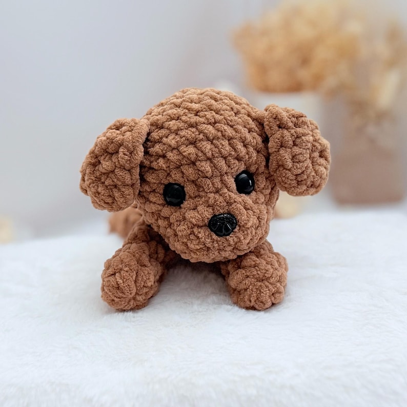 Puede incluir: Un perro de peluche de crochet marr&oacute;n con ojos negros y nariz negra. El perro est&aacute; acostado boca abajo con las patas extendidas.