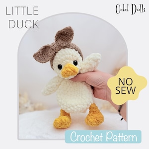 Pode incluir: Um brinquedo de pato de pelúcia crochetado com um laço marrom, corpo branco, bico e pés amarelos. O brinquedo está sendo segurado por uma mão e o texto "NO SEW" é visível no fundo. Os textos "LITTLE DUCK" e "Crochet Pattern" também são visíveis na imagem.