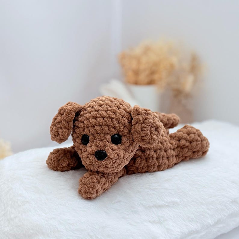 Puede incluir: Un juguete de peluche de perro de crochet marr&oacute;n acostado sobre una superficie blanca y esponjosa.