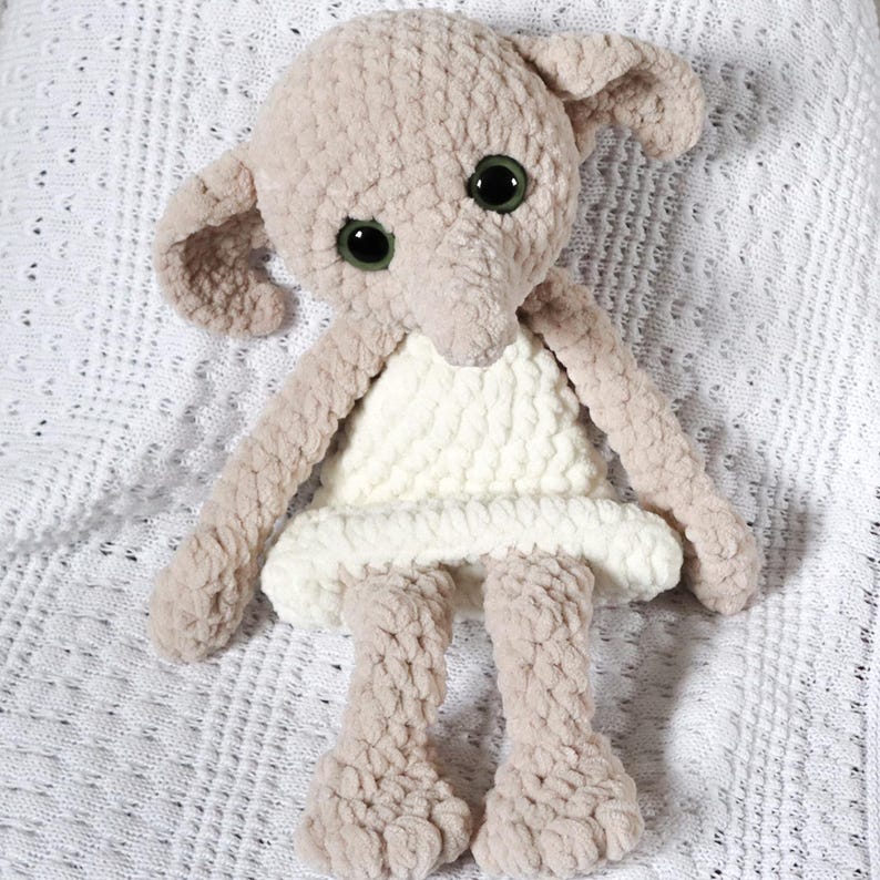 Pode incluir: Um brinquedo de pel&uacute;cia de croch&ecirc; em forma de elfo dom&eacute;stico, com orelhas grandes, um vestido branco e pelo marrom. O brinquedo est&aacute; sentado em um cobertor branco de croch&ecirc;.