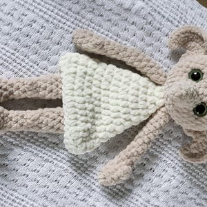 Pode incluir: Um brinquedo de pel&uacute;cia de croch&ecirc; em forma de menina usando um vestido branco. O brinquedo tem um corpo marrom claro e um vestido branco com textura de croch&ecirc;. O brinquedo tem olhos grandes e um nariz pequeno.