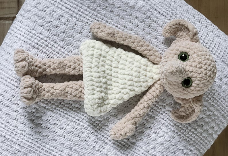 Pode incluir: Um brinquedo de pel&uacute;cia de croch&ecirc; em forma de menina usando um vestido branco. O brinquedo tem um corpo marrom claro e um vestido branco com textura de croch&ecirc;. O brinquedo tem olhos grandes e um nariz pequeno.