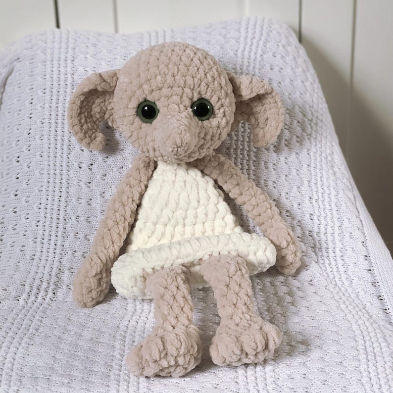 Pode incluir: Um brinquedo de pel&uacute;cia de croch&ecirc; em forma de elfo dom&eacute;stico vestindo um vestido branco. O brinquedo tem grandes olhos castanhos e um nariz pontudo.