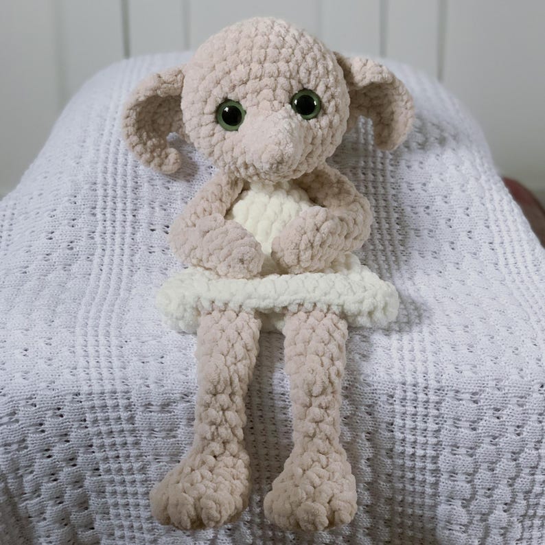 Pode incluir: Um brinquedo de pel&uacute;cia de croch&ecirc; de um elfo dom&eacute;stico vestindo um vestido branco. O elfo tem grandes olhos verdes e um nariz pontudo. O brinquedo est&aacute; sentado em um cobertor de croch&ecirc; branco.