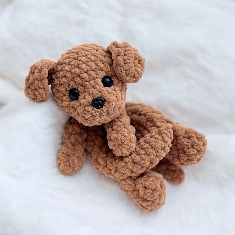 Puede incluir: Un juguete de peluche de perro de crochet marr&oacute;n con ojos negros. El perro est&aacute; acostado sobre una superficie blanca y esponjosa.