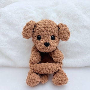 Puede incluir: Un juguete de peluche de perro de crochet marr&oacute;n con ojos negros y nariz negra. El perro est&aacute; sentado con las piernas cruzadas y las patas metidas.