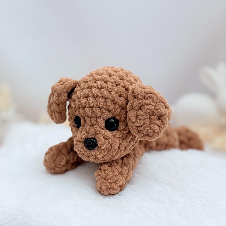 Puede incluir: Un juguete de peluche de caniche de crochet marr&oacute;n con ojos negros y nariz negra. El juguete est&aacute; acostado sobre una superficie blanca y esponjosa.