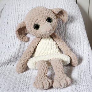 Pode incluir: Um brinquedo de pel&uacute;cia de croch&ecirc; em forma de elfo dom&eacute;stico vestindo um vestido branco. O brinquedo tem grandes olhos verdes e est&aacute; sentado em um cobertor de croch&ecirc; branco.
