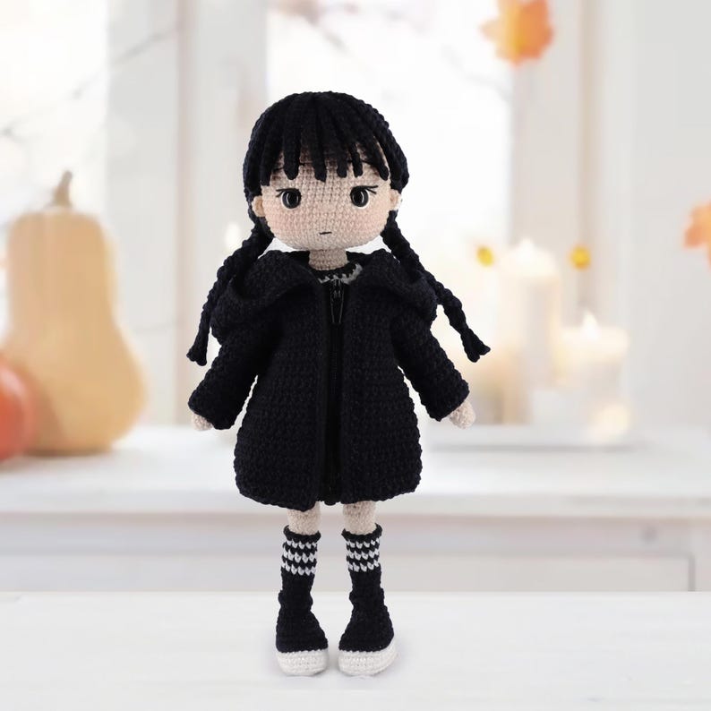 May include: A crocheted doll with black yarn hair styled in two braids and bangs. The doll wears a black hooded jacket, a black dress, and black knee-high socks with white stripes. The doll has a light beige complexion and dark eyes.