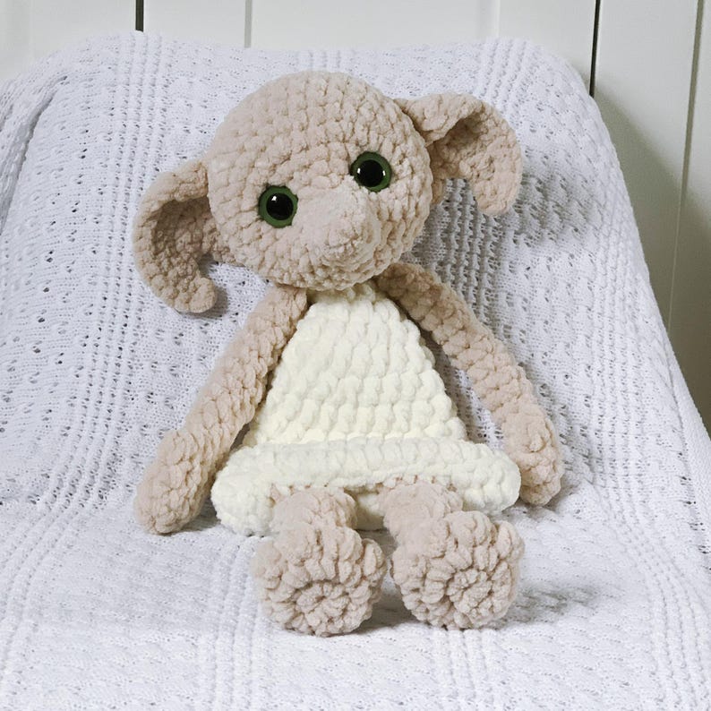 Pode incluir: Um brinquedo de pel&uacute;cia de croch&ecirc; de Dobby, o elfo dom&eacute;stico da s&eacute;rie Harry Potter. O brinquedo est&aacute; vestindo um vestido branco e tem grandes olhos verdes.