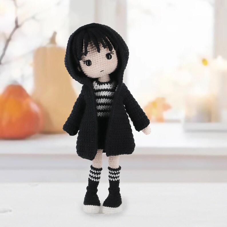 May include: A handmade crochet doll with black hair and a pale complexion, wearing a black hooded cardigan over a black and white striped shirt. The doll features black socks with white stripes and black shoes, crafted with detailed yarn stitching.