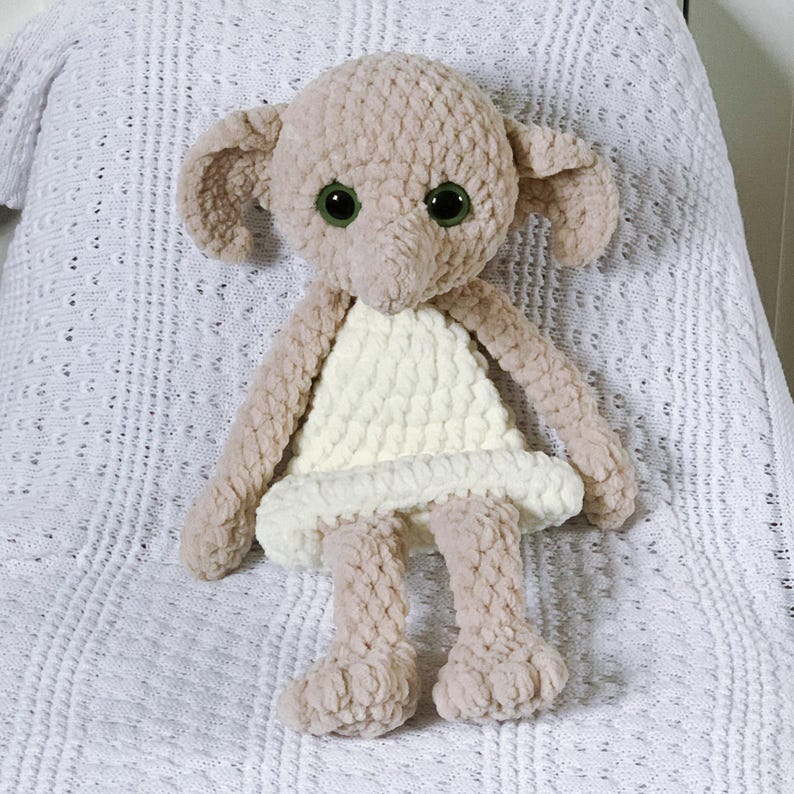 Pode incluir: Um brinquedo de pel&uacute;cia de croch&ecirc; em forma de elfo dom&eacute;stico vestindo um vestido branco. O brinquedo tem grandes olhos verdes e um chap&eacute;u pontudo.