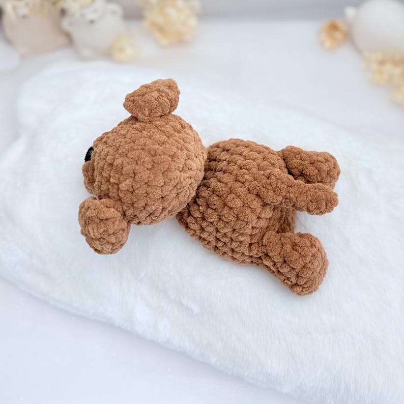 Puede incluir: Un oso de peluche de crochet marr&oacute;n acostado sobre una manta blanca y esponjosa. El oso tiene una peque&ntilde;a nariz negra y ojos.