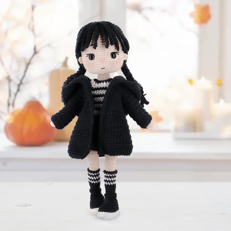 May include: A handmade crochet doll featuring a pale complexion, black hair styled in two braids, and large dark eyes. The doll wears a black hooded cardigan over a black and white striped shirt, a black skirt, and black socks with white stripes.