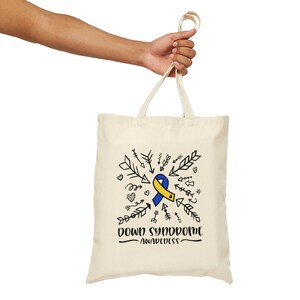 May include: Cream-colored tote bag with a blue and yellow ribbon design, arrows, and the words "DOWN SYNDROME AWARENESS" in black. The bag is held by a person's hand.