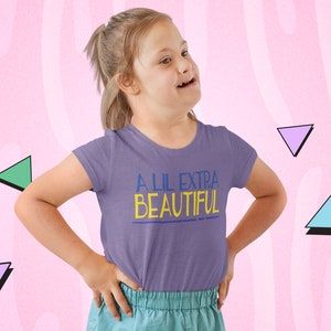 Può includere: Una maglietta viola con la scritta "A LIL EXTRA BEAUTIFUL" in blu e giallo. La maglietta è indossata da una persona con la sindrome di Down. Lo sfondo è rosa con forme geometriche.