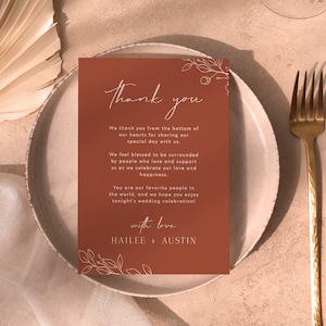 May include: A thank you card with a brown background and white floral illustrations. The text reads "Thank you" in a handwritten font. The card also includes the text "We thank you from the bottom of our hearts for sharing our special day with us. We feel blessed to be surrounded by people who love and support us as we celebrate our love and happiness. You are our favourite people in the world, and we hope you enjoy tonight's wedding celebration! with love HAILEE + AUSTIN"