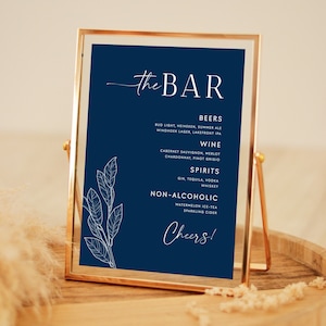 May include: A navy blue sign with white text that reads "The Bar" and lists various alcoholic and non-alcoholic beverages. The sign is framed in a gold metal frame and is sitting on a wooden surface.