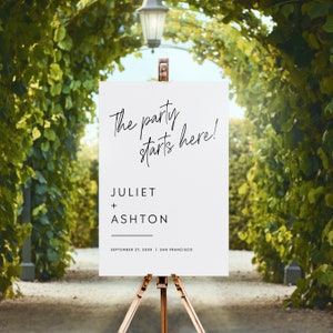 May include: White wedding sign with black calligraphy text that reads "The party starts here!"  The names "Juliet + Ashton" are listed below with the date "September 27, 20XX" and location "San Francisco".