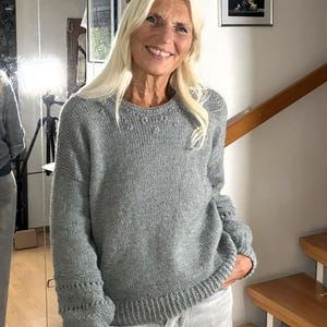 Könnte beinhalten: Ein langärmeliger grauer Strickpullover mit Rundhalsausschnitt, gerippten Bündchen und einem gerippten Saum. Der Pullover weist dezente strukturierte Details um den Kragen sowie horizontale dekorative Stichmuster an den Ärmelbündchen auf.