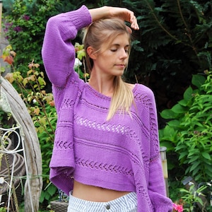 Puede incluir: Un suéter de punto morado con mangas largas y voluminosas y un dobladillo recortado. El suéter presenta detalles calados en el pecho y las mangas. La modelo lleva un pantalón corto a rayas blancas y azules.