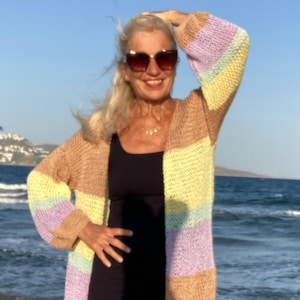 Könnte beinhalten: Ein langer, offener Cardigan in Pastellfarben. Der Pullover hat horizontale Streifen in Beige, Gelb, Lavendel und Hellgrün. Die Ärmel sind lang und der Cardigan wird über einem schwarzen Kleid getragen. Der Hintergrund ist eine Strandszene.