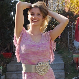 Op de afbeelding: Een roze gebreide jurk zonder mouwen met franjes en een bruine riem met een grote bloemvormige gesp.