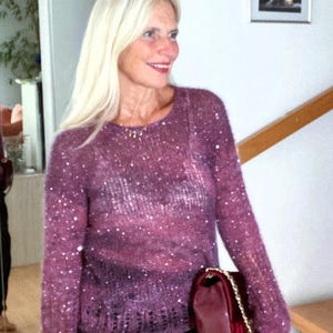 May include: A woman wearing a purple sequined sweater with long sleeves and a round neckline. She is holding a burgundy handbag with a gold chain strap. The sweater is paired with black pants.