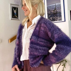 May include: A purple and pink striped cardigan with a fuzzy texture. The cardigan is worn over a white button-down shirt and a purple skirt.