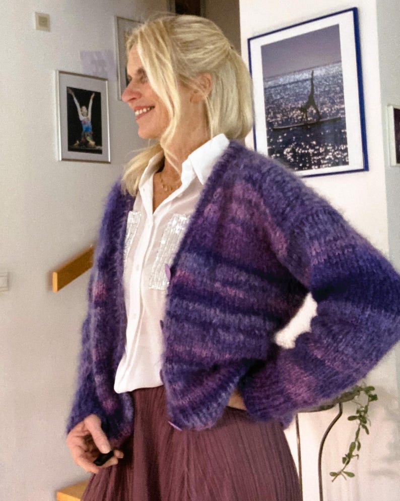 May include: A purple and pink striped cardigan with a fuzzy texture. The cardigan is worn over a white button-down shirt and a purple skirt.
