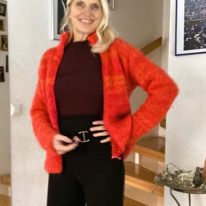 May include: A woman wearing a red and orange striped fuzzy cardigan with a maroon turtleneck shirt and black trousers. She is wearing a gold belt with a large buckle.
