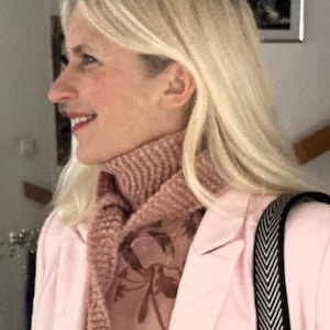May include: A woman wearing a pink blazer with a pink knitted scarf. The scarf is tied in a loose knot at the neck and has a floral design.