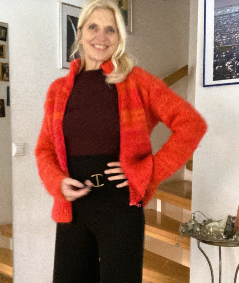 May include: A woman wearing a red and orange striped fuzzy cardigan with a black turtleneck shirt and black trousers. She is wearing a gold belt with a double C buckle.